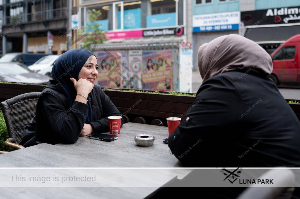 Esra Karamik sitzt draußen an einem Bistrotisch neben einer leicht befahrenen Straße. Auf der anderen Straßenseite reihen sich Läden aneinander. Esra Karamik ist links im Bild und blickt mit einem offenen Lächeln in Richtung einer zweiten Person rechts im Bild an der gegenüberliegenden Seite des Tisches. Beide haben ihre Ellenbogen auf dem Tisch liegen und sind zueinander gewandt. Esra Karamik stützt ihren Kopf auf ihre rechte Hand, am Handgelenk trägt sie einen goldenen Armreif. Sie trägt ein Nasenpiercing, ihre Augen sind leicht geschminkt. Beide Frauen tragen schwarze, langärmelige Oberteile und einen Hijab in dunkelblau bzw. hellbraun. Vor ihnen steht jeweils ein Kaffeebecher, daneben liegen ihre Mobiltelefone.