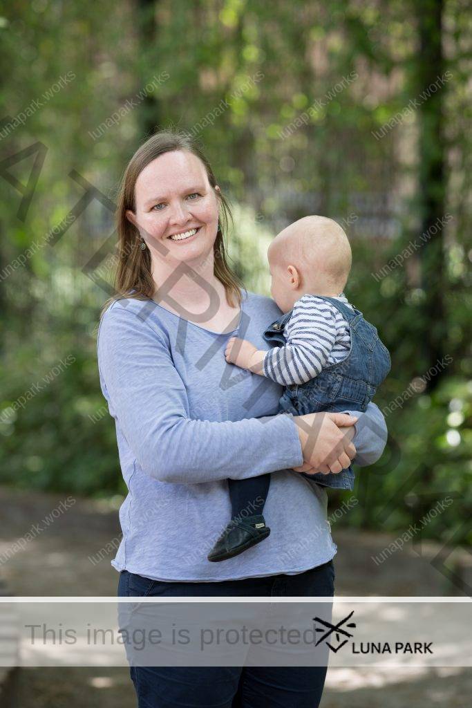 Miriam Stute steht draußen vor einem verschwommenen Hintergrund, der Pflanzen und einen grauen Boden vermuten lässt. Die Sonne scheint. Auf dem Arm hält sie ein Kleinkind, das das Kinn auf ihre linke Schulter legt und in den Bildhintergrund schaut. Miriam Stute blickt mit offenem Lächeln in die Kamera. Sie trägt ein langärmliges, hellblaues T-Shirt und eine dunkelblaue Hose. Ihre schulterlangen, glatten blonden Haare trägt sie offen.