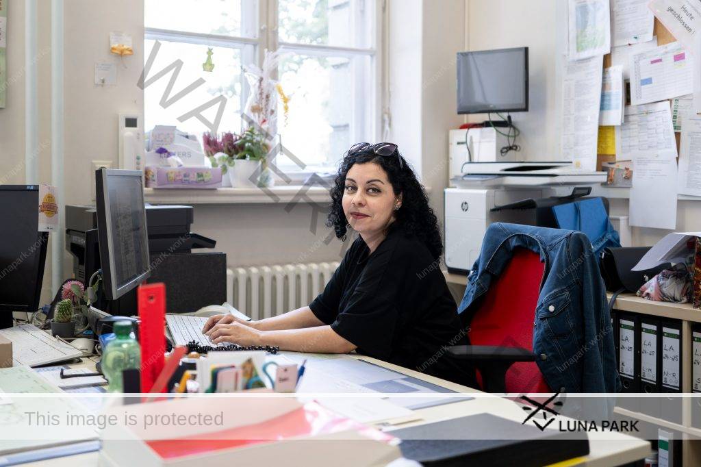 Ebru Schaefer sitzt auf einem Bürostuhl mit roter Lehne an einem großen Schreibtisch vor einem PC. Ihre Unterarme liegen auf dem Tisch ab, ihre Finger berühren eine Computertastatur, ihr Körper ist in Richtung der Tastatur und eines Bildschirms ausgerichtet, ihr Blick geht in Richtung Kamera, sie lächelt leicht. Auf dem Schreibtisch sind viele Büromaterialien, links im Bild ist ein weiterer Arbeitsplatz zu erkennen. Im Bildhintergrund ist eine Pinnwand voller bedruckter Zettel, ein kleines Regal, in dem Aktenordner stehen,  ist durch den Bürostuhl teils verdeckt. Auf dem Regal liegen eine schwarze Handtasche, ein blauer Stoffbeutel und ein Schal. Ebru Schäfer trägt ein schwarzes, weites, kurzärmliges Tshirt, ihre schwarzen, lockigen Haare liegen offen und gehen etwas über die Schulter, eine Sonnenbrille hält sie wie ein Haarreif aus dem Gesicht. An ihrem linken Ohr trägt sie einen  silbernen Ohrring und einen Piercing, der weiß glitzert. Ihre Augen sind geschminkt, sie trägt Lippenstift.