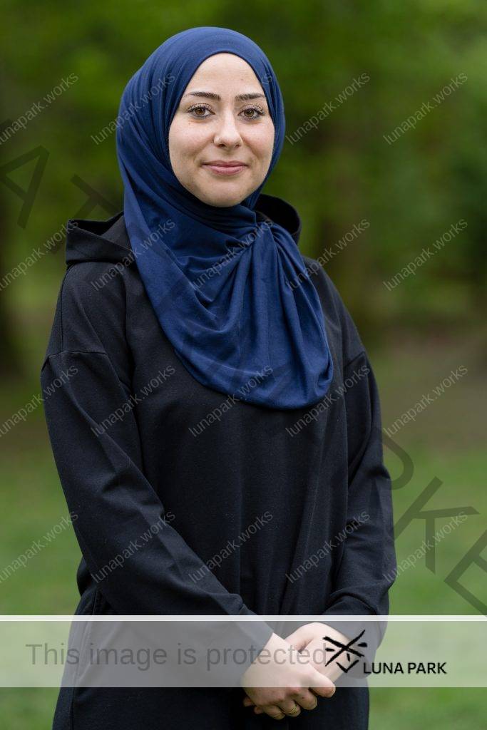 Esra Karamik sitzt draußen an einem Bistrotisch neben einer leicht befahrenen Straße. Auf der anderen Straßenseite reihen sich Läden aneinander. Esra Karamik ist links im Bild und blickt mit einem offenen Lächeln in Richtung einer zweiten Person rechts im Bild an der gegenüberliegenden Seite des Tisches. Beide haben ihre Ellenbogen auf dem Tisch liegen und sind zueinander gewandt. Esra Karamik stützt ihren Kopf auf ihre rechte Hand, am Handgelenk trägt sie einen goldenen Armreif. Sie trägt ein Nasenpiercing, ihre Augen sind leicht geschminkt. Beide Frauen tragen schwarze, langärmelige Oberteile und einen Hijab in dunkelblau bzw. hellbraun. Vor ihnen steht jeweils ein Kaffeebecher, daneben liegen ihre Mobiltelefone.