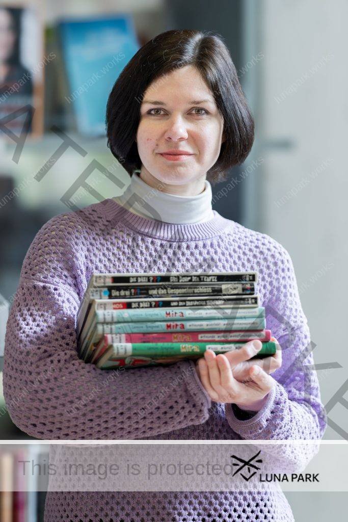 Nadjeschda Pugatschowa steht vor einem Bücherregal, das verschwommen zu erkennen ist. Sie blickt mit einem leichten Lächeln in die Kamera. Ihr rechter Arm ist vor ihrem Körper angewinkelt und wird von ihrem linken Arm gestützt. So hält sie einen Bücherstapel aus 3 Büchern von “Die drei Fragezeichen” und 4 Büchern aus einer Reihe mit dem Titel “Mira”. Die Bücher tragen Kennzeichnungen einer Bibliothek. Ihre glatten, dunkelbraunen Haare trägt sie in einem Kurzhaarschnitt bis zum Kinn, unter ihrem langärmligen, fliederfarbenen Pullover trägt sie ein weißes Rollkragenshirt.