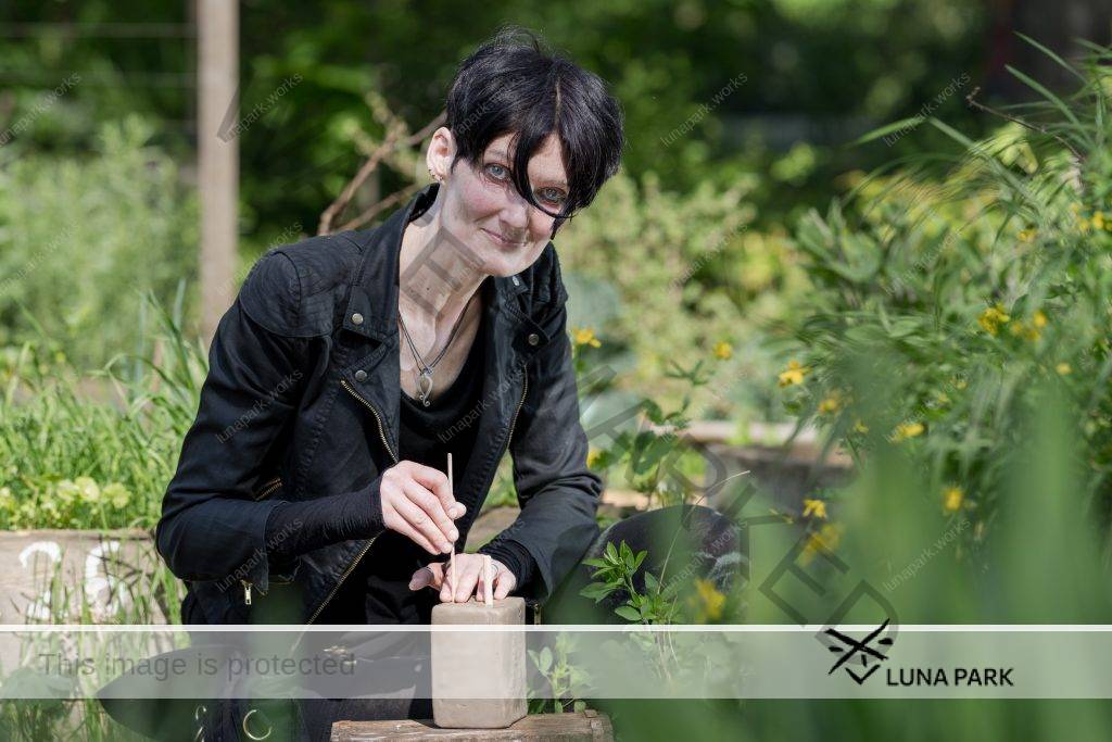 Jeanine Fornaçon kniet draußen zwischen Blumen und Gräsern, die teils in Kisten wachsen. Vor ihr liegt auf einem Holzblock ein Klumpen Ton, den sie mit zwei kleinen Holzstäben bearbeitet. Sie blickt mit einem Lächeln in die Kamera, eine schwarze Haarsträhne fällt aus ihrem Kurzhaarschnitt ins Gesicht. Über einem schwarzen Tshirt trägt sie offen eine Bikerjacke aus schwarzem Stoff. Sie trägt eine schwarze kurze Hose und schwarze Leggings. Um ihren Hals trägt sie an einem dünnen Lederband einen silbernen Anhänger